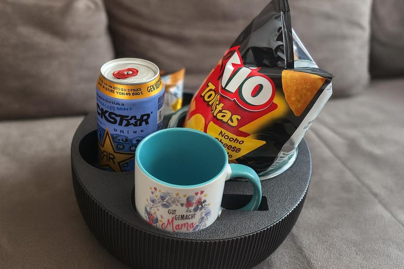 May include: A black, round, multi-compartment tray holds a can of Rockstar energy drink, a bag of Tio Nacho cheese tortilla chips, and a white mug with a blue handle and interior. The mug has floral and text designs.