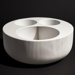 Può includere: Una ciotola decorativa bianca e circolare con tre aperture circolari più piccole e una grande apertura centrale. L'esterno presenta dettagli verticali e scanalati. La ciotola è posta su uno sfondo nero.