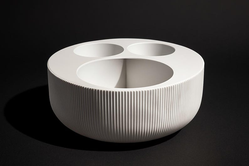 May include: A white, circular decorative bowl with three smaller circular openings and a large central opening. The exterior features vertical, ridged detailing. The bowl is set against a black background.