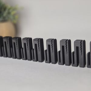 Puede incluir: Una fila de clips negros rectangulares con una abertura en forma de U y un borde en zigzag. Los clips están dispuestos sobre una superficie blanca, con un fondo borroso.
