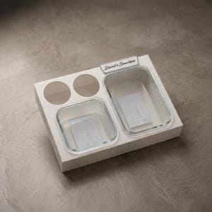 Puede incluir: Una bandeja de cartón blanco para aperitivos con dos recortes redondos y dos recipientes de vidrio transparente. La bandeja tiene el texto "David's Snackbar" impreso. Los recipientes son rectangulares y cuadrados.