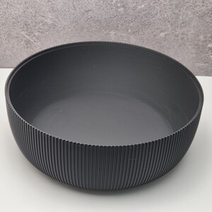 May include: A dark gray bowl with a ribbed exterior. The bowl has a wide, shallow design and a smooth interior. The vertical ribbing on the outside adds texture. The bowl is likely made of a durable material.