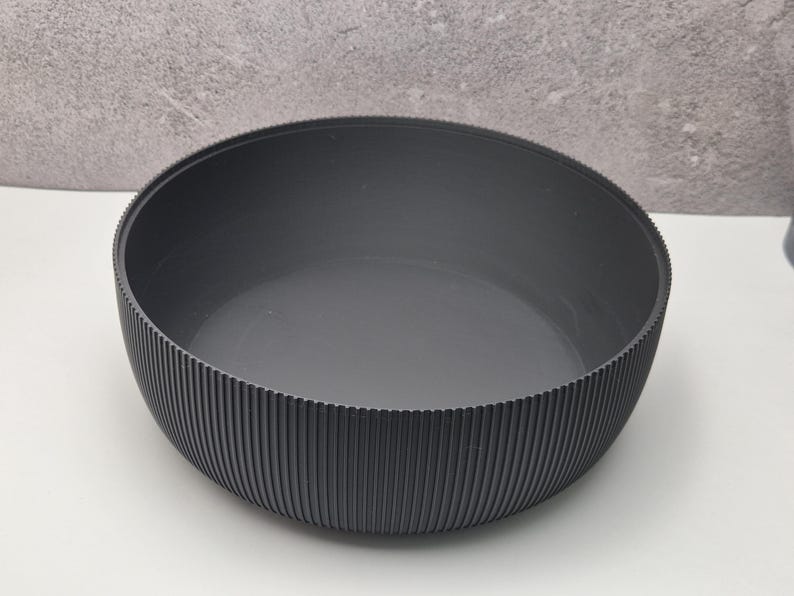 May include: A dark gray bowl with a ribbed exterior. The bowl has a wide, shallow design and a smooth interior. The vertical ribbing on the outside adds texture. The bowl is likely made of a durable material.