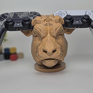 May include: A wooden bull head-shaped gaming controller stand. The stand holds two controllers, one with a camouflage design and the other white. The bull head is intricately carved with detailed lines, and the stand is on a round base.