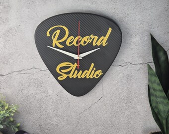 May include: Black guitar pick-shaped wall clock with the words "Record Studio" in gold script. The clock has white hands and a red second hand. The background is a textured gray wall with a few plants.