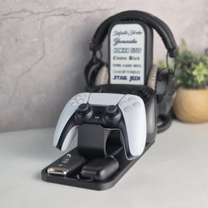 Könnte beinhalten: Schwarzer Gaming-Konsolenständer mit einem weiß-schwarzen Controller, Kopfhörern und einer Ladestation. Der Ständer trägt den Text "Infinite Stroke, Yanaresha, GEMZA 7770, Cluster Black, PAPER DASH BOSS, GAME OF THRONES, STAR JEDI."