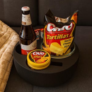 May include: A black, round serving tray holds a bottle of beer, a bag of tortilla chips, and a container of dip. The tortilla chips are labeled "Nacho Cheese Style." The tray is on a gray surface.