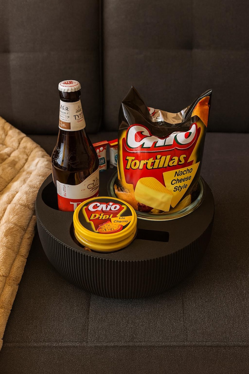 May include: A black, round serving tray holds a bottle of beer, a bag of tortilla chips, and a container of dip. The tortilla chips are labeled "Nacho Cheese Style." The tray is on a gray surface.
