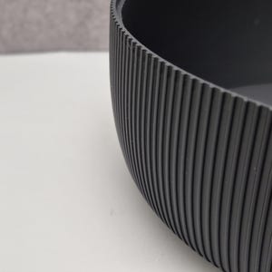 May include: A close-up of a dark gray, oval-shaped bowl. The exterior features vertical, ridged detailing. The bowl is set against a white surface with a gray background.