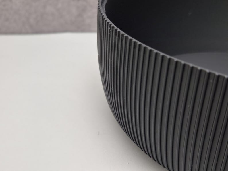 May include: A close-up of a dark gray, oval-shaped bowl. The exterior features vertical, ridged detailing. The bowl is set against a white surface with a gray background.