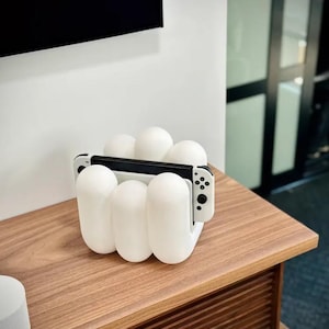 Könnte beinhalten: Eine weiße Nintendo Switch-Konsole und Joy-Con-Controller, die in einem weißen, wolkenförmigen Ständer auf einer Holzoberfläche liegen.