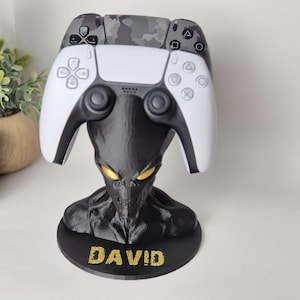 May include: A black and white alien-themed controller stand with the name "DAVID" in gold lettering. The stand holds a white and camouflage-patterned game controller. The alien has glowing yellow eyes.