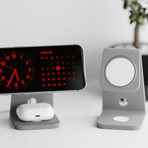 Könnte beinhalten: Ein grauer Telefonständer mit einem schwarzen Telefon, das eine rote Digitaluhr und einen Kalender anzeigt. Das Telefon steht auf einer weißen Oberfläche. Rechts im Bild befindet sich ein grauer Ständer für eine Uhr oder ein anderes Gerät.