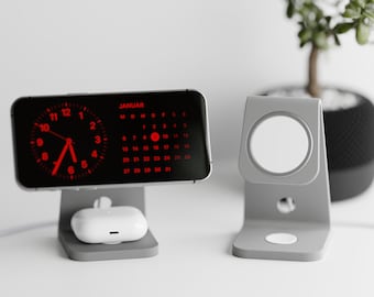 May include: A gray phone stand with a black phone displaying a red digital clock and calendar. The phone is on a white surface. There is a gray stand for a watch or other device on the right side of the image.