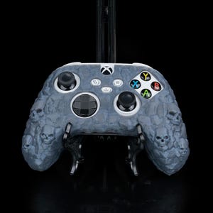 May include: A gray and black Xbox controller with a skull pattern. The controller is on a black stand.