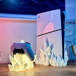 Könnte beinhalten: Eine weiße PlayStation 5 Konsole mit einem kristallförmigen Ständer. Ein weißer kristallförmiger Ständer mit einem blauen PlayStation 5 Controller darauf. Die Konsole und der Controller werden mit einem warmweißen Licht beleuchtet.