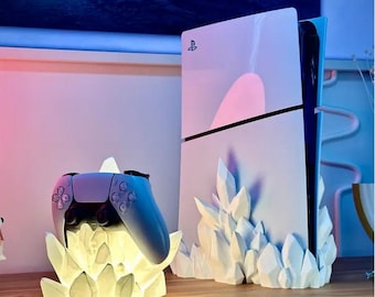 May include: A white PlayStation 5 console with a crystal-shaped stand. A white crystal-shaped stand with a blue PlayStation 5 controller on top. The console and controller are illuminated with a warm white light.