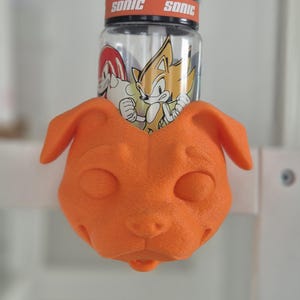 Könnte beinhalten: Ein orangefarbener, 3D-gedruckter Hundehalter mit einer transparenten Wasserflasche mit Sonic the Hedgehog-Grafiken. Die Flasche hat einen schwarzen Deckel mit orangefarbenem "SONIC"-Aufdruck. Der Halter ist auf einer weißen Oberfläche montiert.