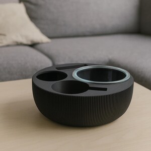 May include: A black, round, multi-functional tabletop organizer with a ribbed exterior. It features three circular compartments, including a clear glass bowl. The organizer sits on a light-colored wooden surface, with a gray sofa and pillow in the background.