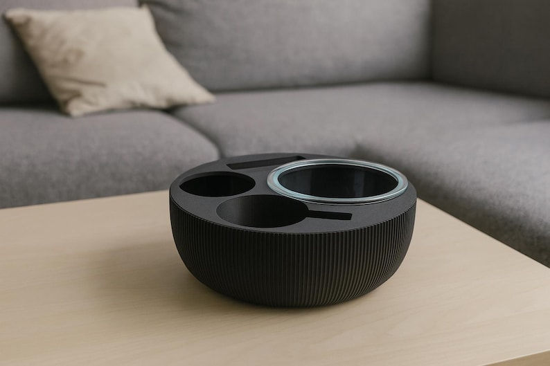 May include: A black, round, multi-functional tabletop organizer with a ribbed exterior. It features three circular compartments, including a clear glass bowl. The organizer sits on a light-colored wooden surface, with a gray sofa and pillow in the background.
