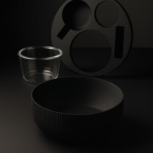 May include: A still-life composition featuring a black bowl with vertical ridges, a clear glass, and a circular black tray with cut-out shapes. The objects are set against a dark background, creating a minimalist aesthetic.