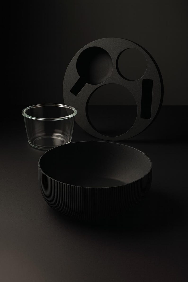 May include: A still-life composition featuring a black bowl with vertical ridges, a clear glass, and a circular black tray with cut-out shapes. The objects are set against a dark background, creating a minimalist aesthetic.