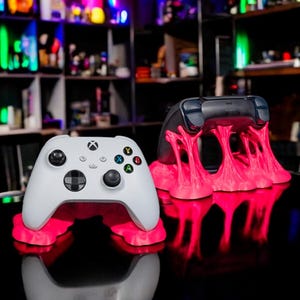 Könnte beinhalten: Zwei weiße Videospiel-Controller auf rosa, 3D-gedruckten Ständern. Die Controller befinden sich auf einer schwarzen Oberfläche mit einer Reflexion der Controller und Ständer.