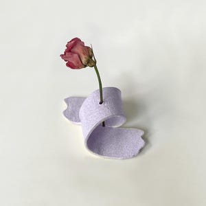 Pode incluir: Um vaso de cerâmica lavanda em forma de fita segura uma rosa rosa seca. O vaso tem uma superfície texturizada e um pequeno orifício para segurar o caule da flor. O fundo é branco liso.
