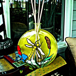 Könnte beinhalten: Eine klare Glasdiffuserflasche mit einem gelben, grünen und weißen Libellen-Design. Die Flasche hat Holzstäbchen im Inneren und steht auf einem Tisch mit einem schwarz-weiß karierten Muster.