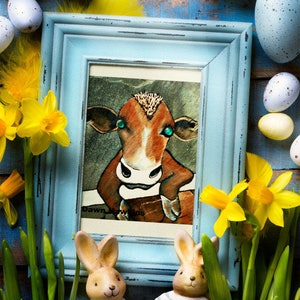 May include: A framed illustration of a brown cow with green eyes, surrounded by Easter eggs, daffodils, and two bunny figurines. The frame is light blue and the illustration is titled "Dawn Meadows".