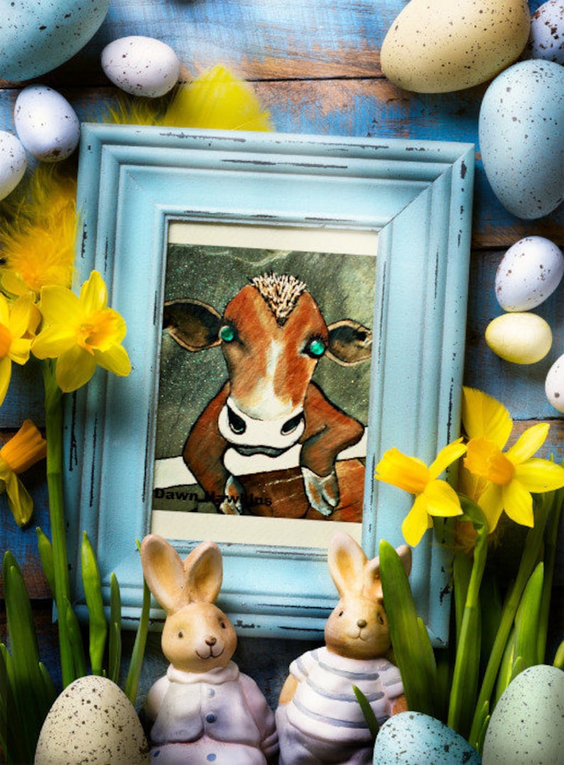 May include: A framed illustration of a brown cow with green eyes, surrounded by Easter eggs, daffodils, and two bunny figurines. The frame is light blue and the illustration is titled "Dawn Meadows".