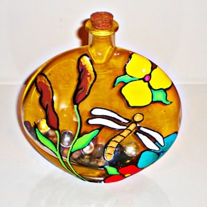 Könnte beinhalten: Eine runde Glasflasche mit einem Korkverschluss, bemalt mit einer Libelle, Blumen und Schilf. Die Flasche ist mit kleinen, klaren Glasperlen gefüllt.
