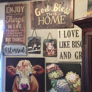 May include: A collection of framed wall art with various themes, including a cow, flowers, and inspirational quotes. The quotes include "Enjoy the little things in life", "God bless this home", "I love like biscuits and gravy", and "Blessed".