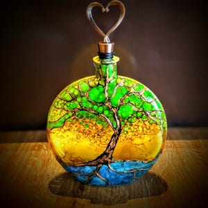 May include: A decorative glass bottle with a heart-shaped stopper. The bottle features a painted tree design with green, yellow, and blue colors. The tree's branches and trunk are outlined in gold. The bottle is sitting on a wooden surface.