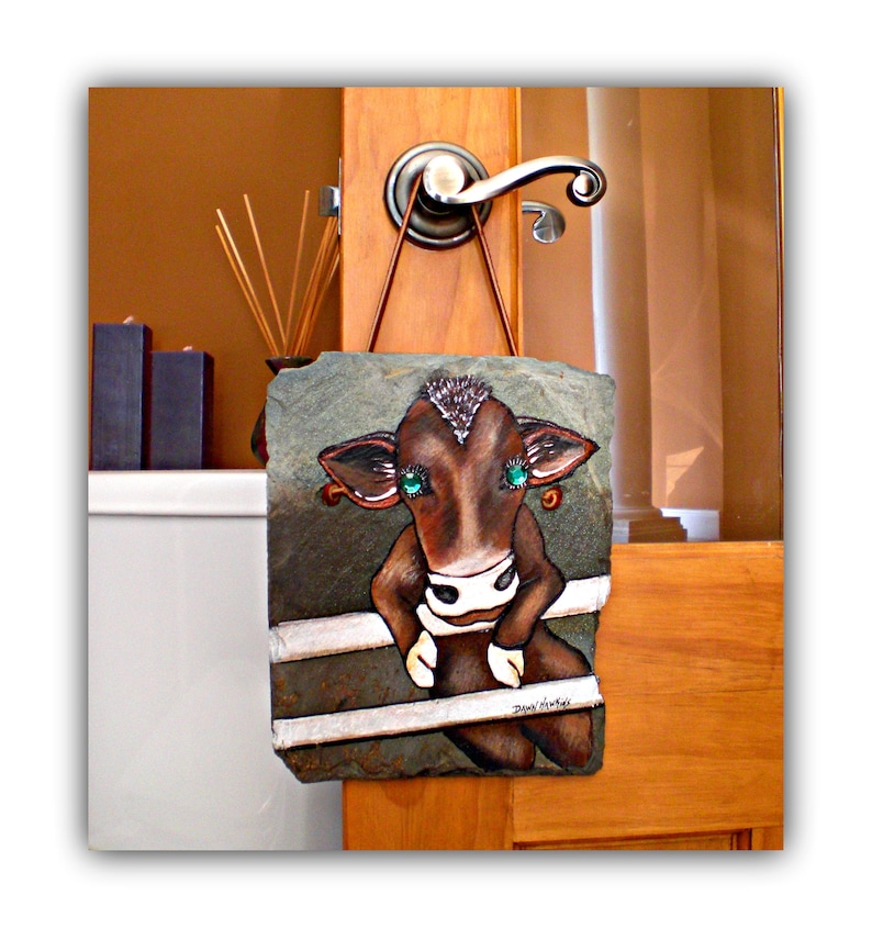 May include: A brown cow with green eyes painted on a slate plaque. The plaque is hanging on a wooden door with a silver door handle.