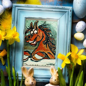 May include: A blue picture frame with a cartoon horse illustration inside. The frame is surrounded by Easter eggs, daffodils, and two bunny figurines.