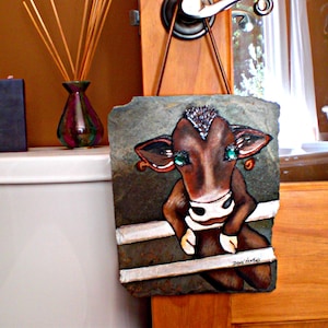 May include: A brown and white cow with green eyes painted on a slate plaque. The plaque is hanging on a wooden door. The cow is looking at the viewer. The plaque is signed "Dawn Marie".