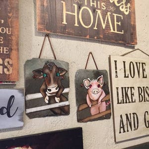 May include: Two painted slate signs with a cow and a pig on them. The cow is brown with green eyes and the pig is pink with a red nose. Both animals are standing in a wooden fence. The signs are hanging on a wall with other signs that say "God Bless This Home" and "I Love Like Biscuits and Gravy".