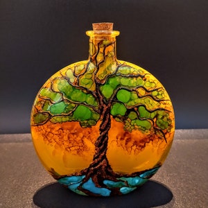 May include: A round glass bottle with a cork stopper, hand-painted with a tree of life design. The tree has a brown trunk, green leaves, and blue roots. The background is a yellow-orange color.