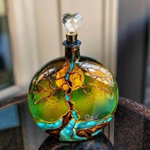 Könnte beinhalten: Eine dekorative Glasflasche mit herzförmigem Verschluss. Die Flasche hat ein Baumdesign mit grünen, gelben und braunen Farben. Das Wort "Dad" ist in Weiß auf die Flasche geschrieben. Die Flasche ist rund und hat einen goldfarbenen Rand.