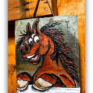 May include: A hand-painted slate sign with a cartoon horse with brown fur, a black mane, and a white face. The horse is smiling with its mouth open and has large, round eyes. The sign is hanging from a leather strap and has the word "WILLOW" written on it in white paint. The sign is signed "DAWN HAWKINS" in the bottom right corner.