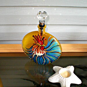 Può includere: Una bottiglia di vetro gialla con un tappo di cristallo, dipinta a mano con un motivo di gallo colorato. La bottiglia è su una superficie di legno.