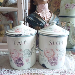 Pode incluir: Dois recipientes cilíndricos de esmalte na cor creme com tampas a condizer, cada um com um padrão floral de rosas em tons de rosa e roxo. O recipiente da esquerda tem a palavra CAFÉ e o da direita tem a palavra SUCRE.