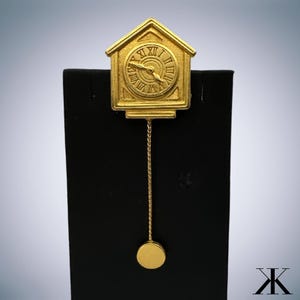 May include: A gold-colored cuckoo clock brooch with a pendulum. The clock face has Roman numerals and a black hand.