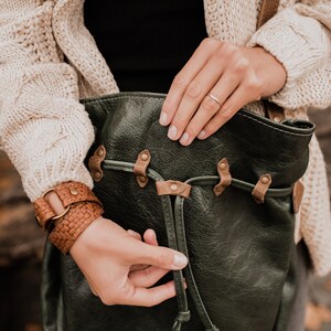 May include: A dark green leather bag with brown leather straps and metal accents. The bag has a drawstring closure and is being held by a person.