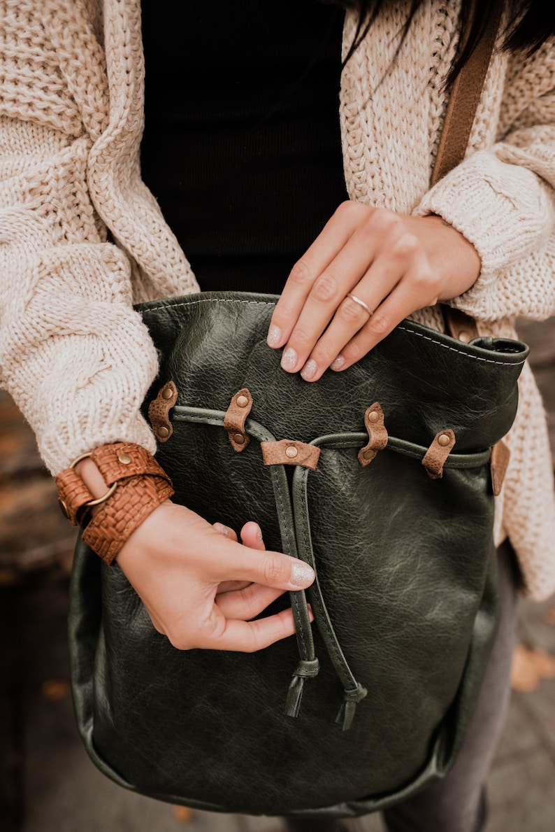 May include: A dark green leather bag with brown leather straps and metal accents. The bag has a drawstring closure and is being held by a person.