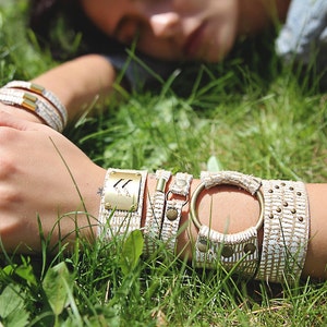 May include: A close-up of a person's wrist wearing several bracelets. The bracelets are made of white woven fabric with gold accents. The bracelets are layered on top of each other and have a variety of textures.