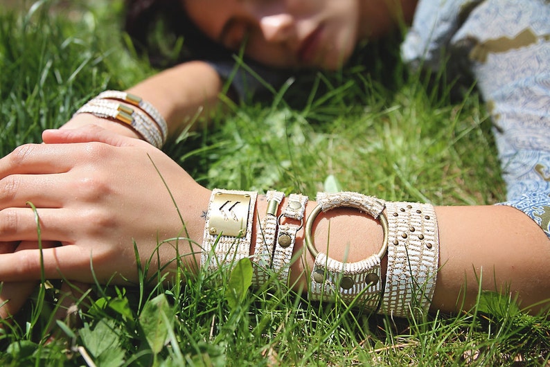 May include: A close-up of a person's wrist wearing several bracelets. The bracelets are made of white woven fabric with gold accents. The bracelets are layered on top of each other and have a variety of textures.