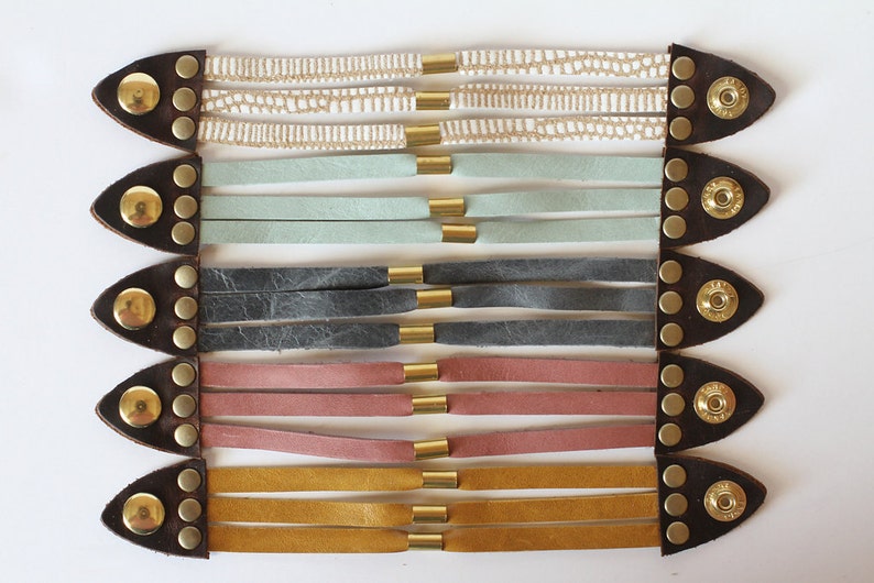 May include: A set of six leather bracelets with gold accents. The bracelets are layered in a rainbow pattern, starting with a white and cream bracelet, followed by a light blue bracelet, a grey bracelet, a pink bracelet, and a yellow bracelet.