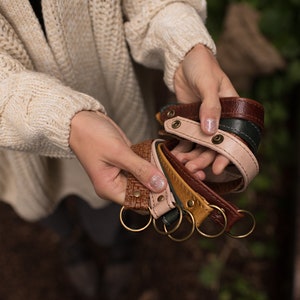 May include: A set of five leather keychains in various colors, including brown, pink, green, and yellow. Each keychain has a metal ring and a decorative stitch detail.
